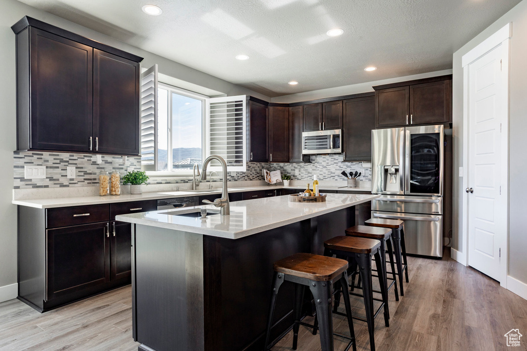Kitchen featuring an island with sink, light wood-type flooring, stainless steel appliances, a breakfast bar area, and backsplash