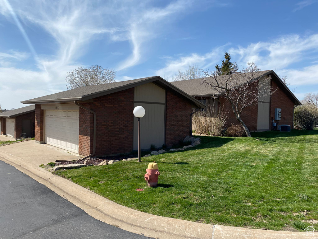 View of side of property featuring central air condition unit, driveway, brick siding, a garage, and a lawn