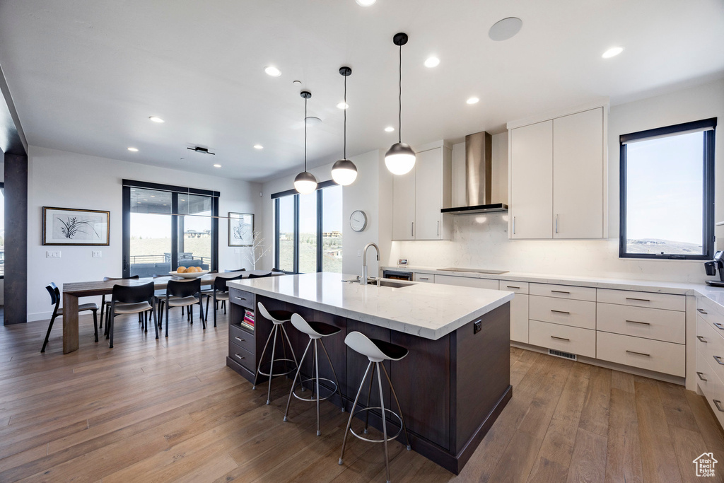Kitchen featuring white cabinets, a center island with sink, wall chimney exhaust hood, hardwood / wood-style flooring, and a sink