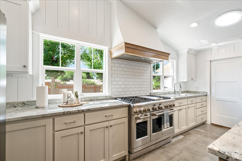 Kitchen with custom exhaust hood, tasteful backsplash, vaulted ceiling, light stone countertops, and range with two ovens