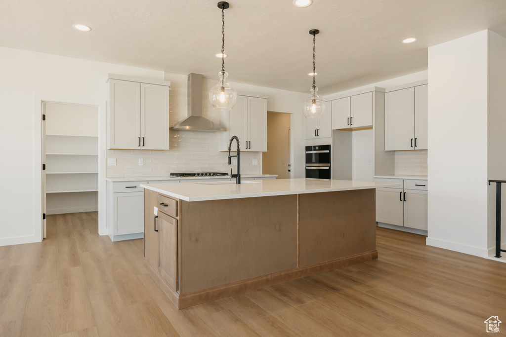 Kitchen featuring wall chimney range hood, tasteful backsplash, a center island with sink, light wood finished floors, and recessed lighting