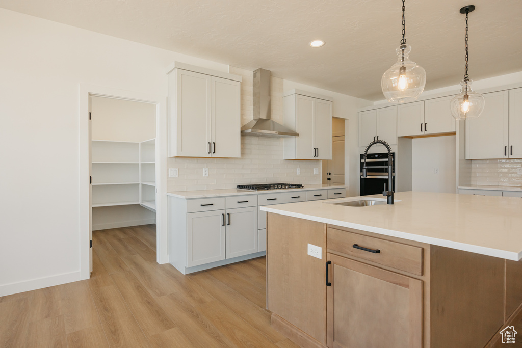 Kitchen featuring wall chimney exhaust hood, a sink, light wood finished floors, decorative backsplash, and light countertops