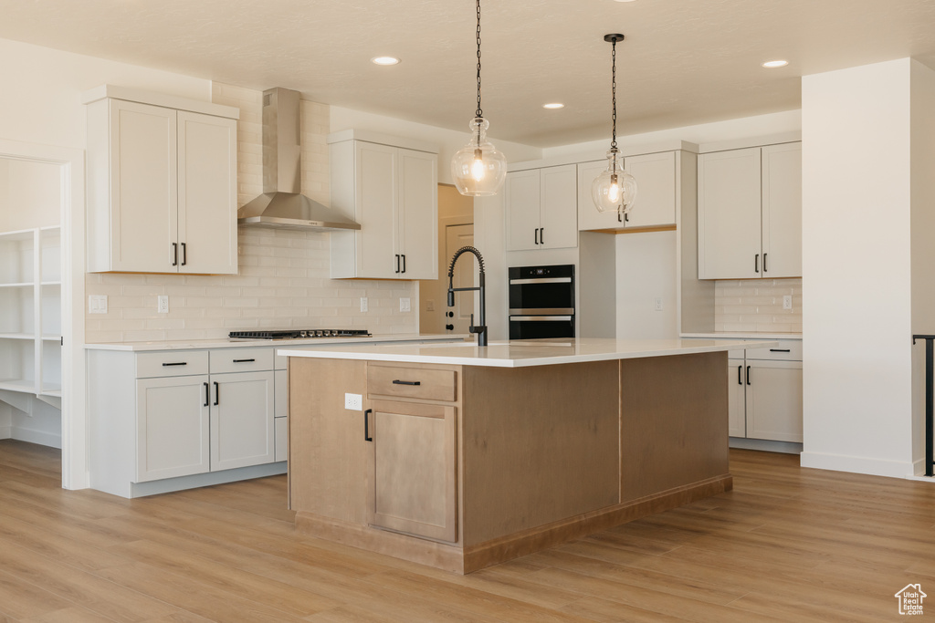 Kitchen featuring wall chimney exhaust hood, tasteful backsplash, light wood finished floors, a kitchen island with sink, and recessed lighting