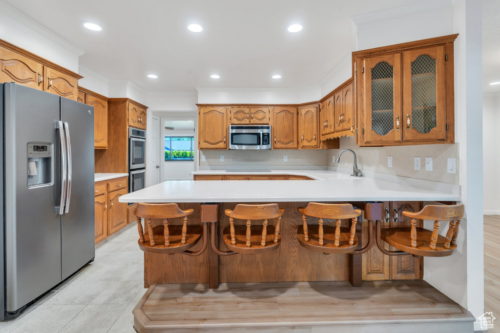 Kitchen featuring stainless steel appliances, a peninsula, light countertops, glass insert cabinets, and brown cabinets