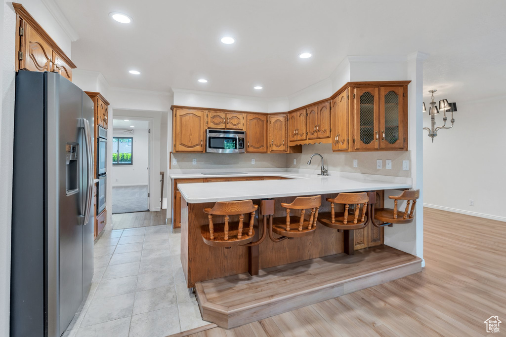Kitchen featuring appliances with stainless steel finishes, a peninsula, light countertops, brown cabinetry, and glass insert cabinets