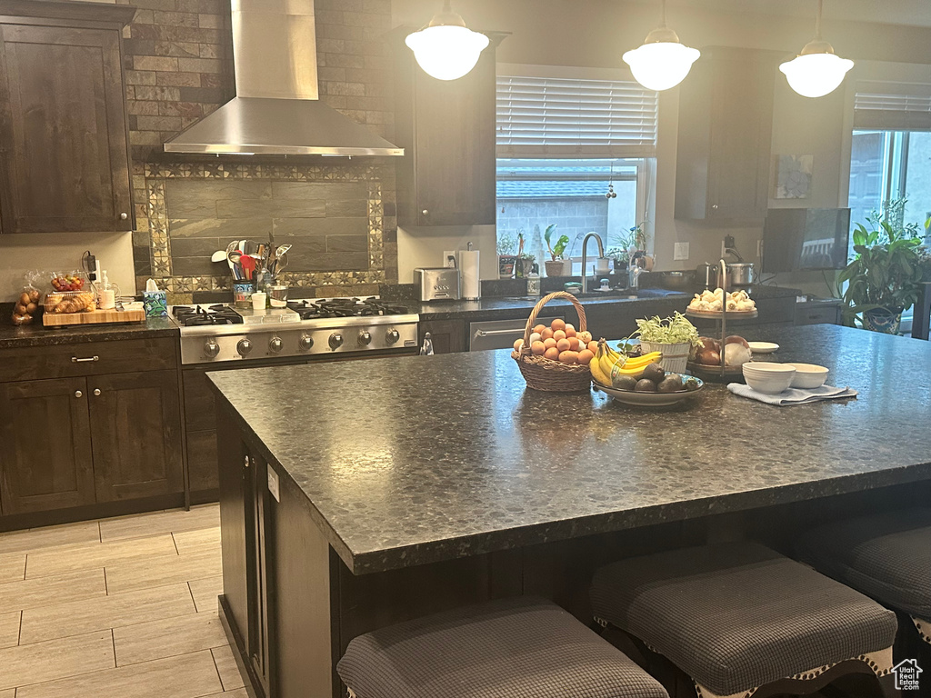 Kitchen with dark brown cabinets, decorative light fixtures, wall chimney exhaust hood, stainless steel gas cooktop, and a breakfast bar area