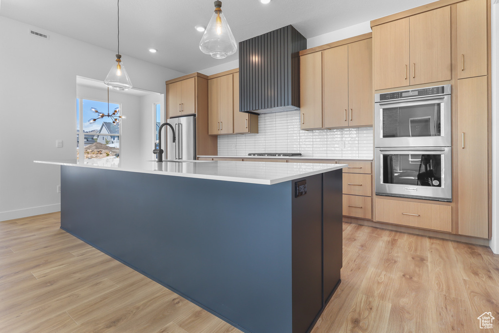 Kitchen with appliances with stainless steel finishes, a kitchen island with sink, light countertops, light brown cabinetry, and backsplash
