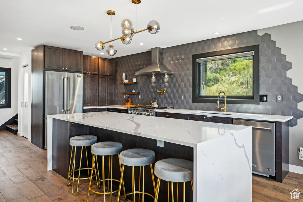 Kitchen featuring dark brown cabinetry, wall chimney range hood, light stone countertops, stainless steel appliances, and a sink