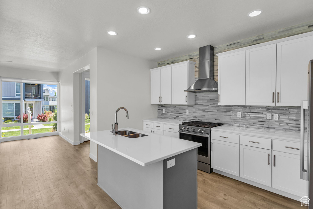 Kitchen featuring wall chimney exhaust hood, stainless steel range with gas cooktop, a sink, backsplash, and a kitchen island with sink