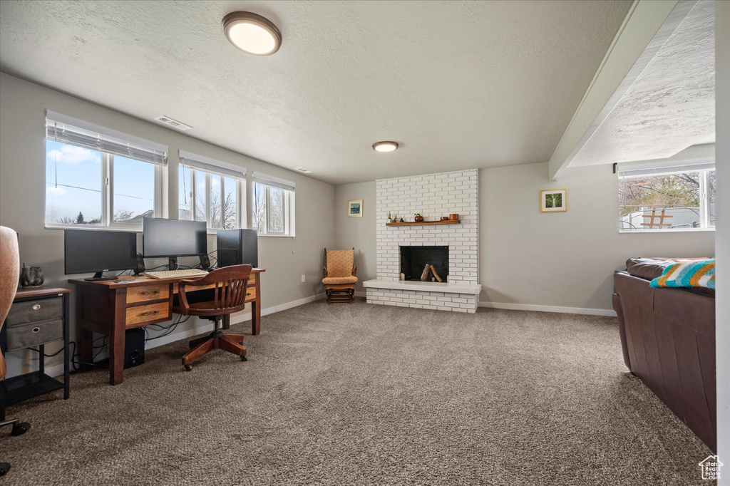 Carpeted home office with a brick fireplace, a textured ceiling, baseboards, and visible vents