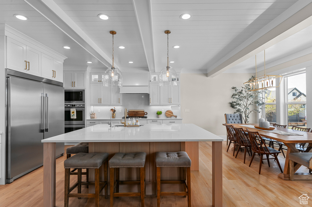 Kitchen with built in appliances, light countertops, beamed ceiling, white cabinetry, and light wood finished floors