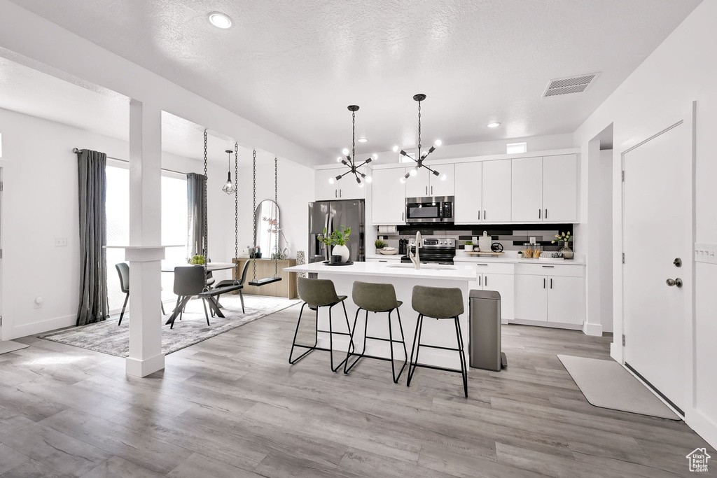 Kitchen featuring stainless steel appliances, a chandelier, an island with sink, white cabinets, and a kitchen breakfast bar