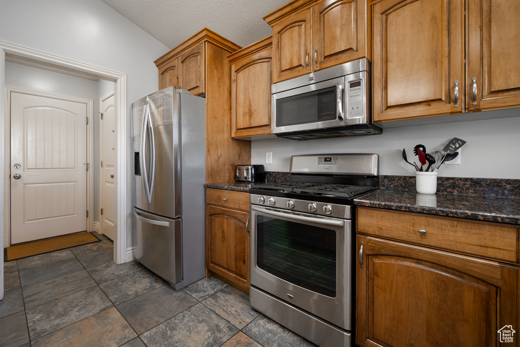 Kitchen featuring dark stone countertops, brown cabinetry, stone finish floor, and stainless steel appliances