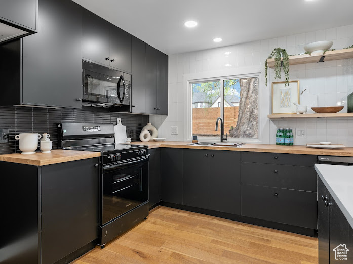 Kitchen featuring dark cabinetry, a sink, black appliances, wood counters, and light wood-style flooring