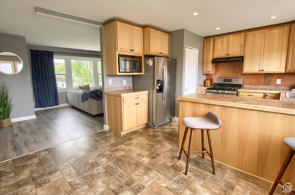 Kitchen featuring stainless steel appliances, stone finish flooring, light countertops, under cabinet range hood, and a breakfast bar