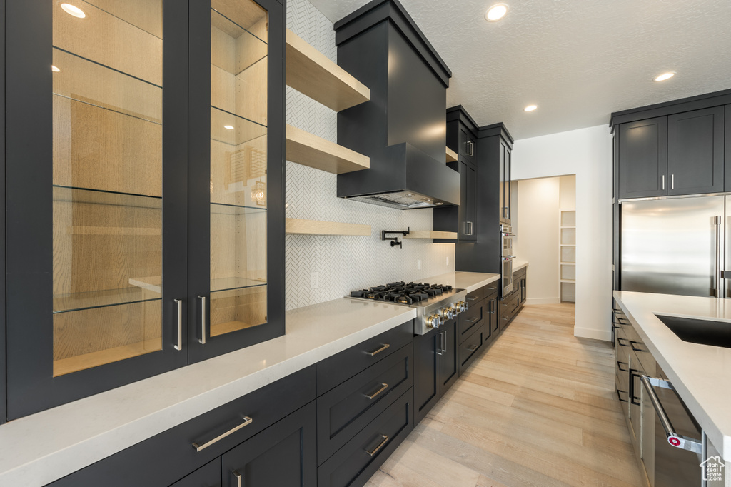 Kitchen featuring appliances with stainless steel finishes, wall chimney exhaust hood, light wood-style flooring, dark cabinetry, and open shelves