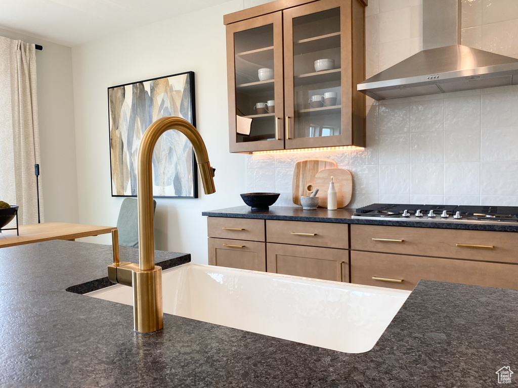 Kitchen with backsplash, wall chimney range hood, a sink, stainless steel gas cooktop, and glass insert cabinets