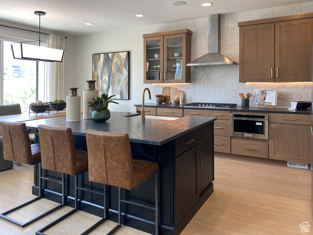 Kitchen with wall chimney exhaust hood, backsplash, glass insert cabinets, a breakfast bar, and appliances with stainless steel finishes