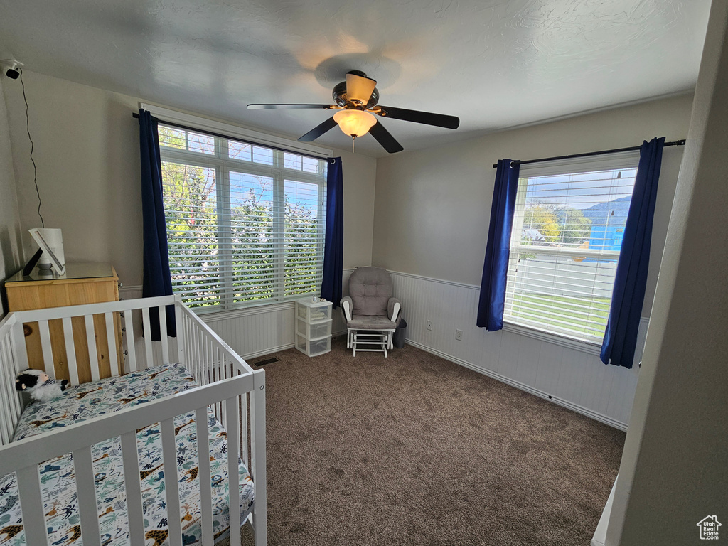 Carpeted bedroom featuring a nursery area, multiple windows, a ceiling fan, and wainscoting