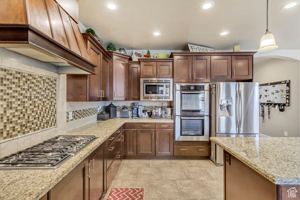 Kitchen with decorative backsplash, stainless steel appliances, premium range hood, light stone countertops, and decorative light fixtures