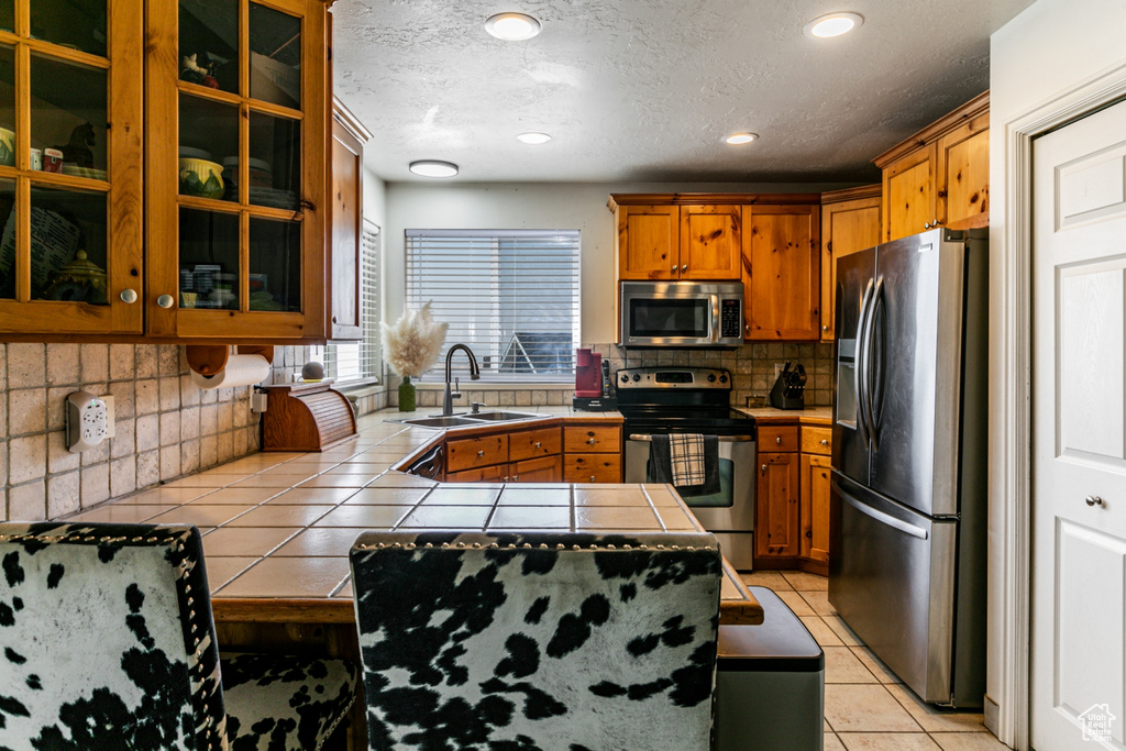 Kitchen featuring brown cabinets, tile countertops, a sink, decorative backsplash, and appliances with stainless steel finishes