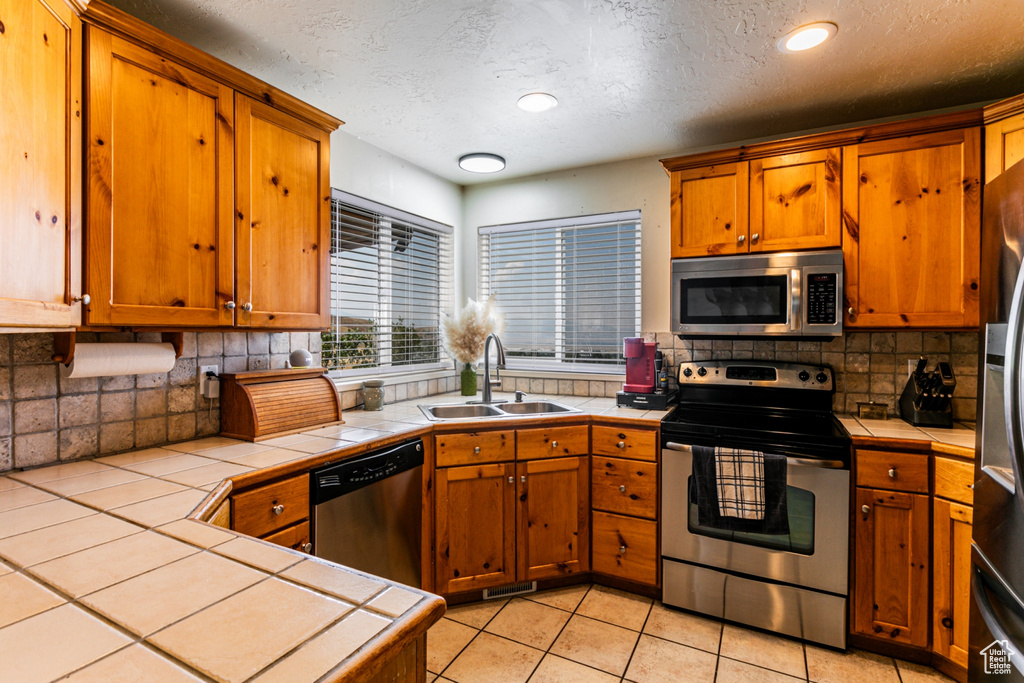 Kitchen with a textured ceiling, a sink, brown cabinets, tile counters, and appliances with stainless steel finishes