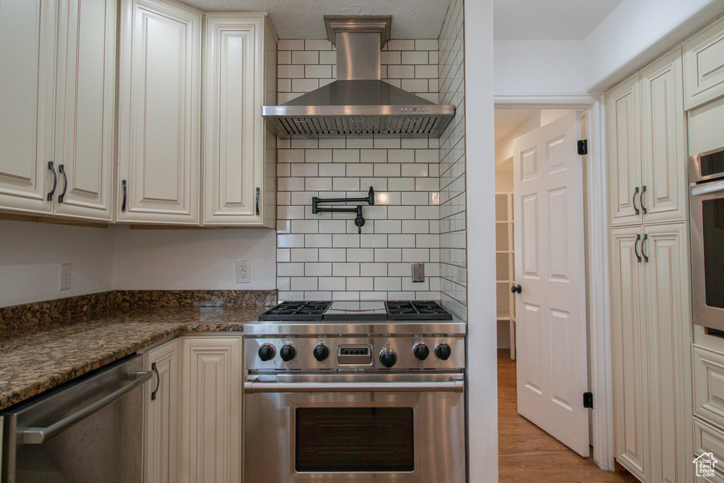 Kitchen featuring wall chimney exhaust hood, stainless steel appliances, dark stone counters, backsplash, and light wood-style floors