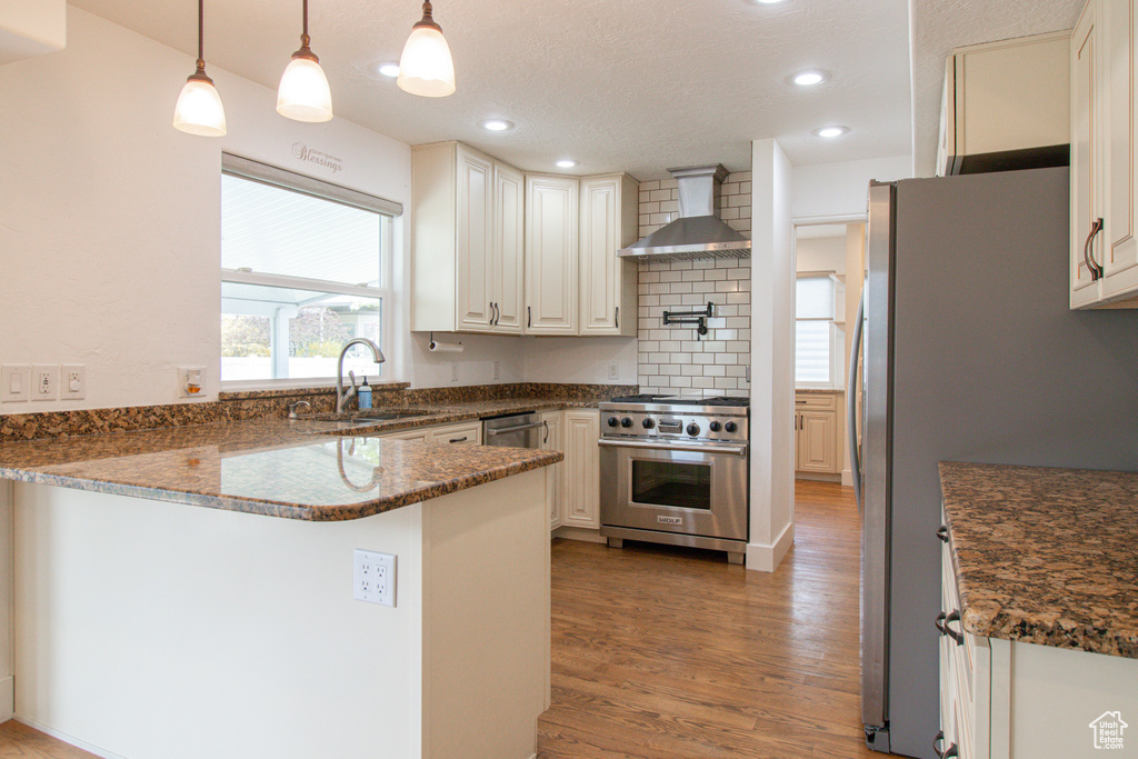 Kitchen with wall chimney range hood, a sink, light wood-style floors, a peninsula, and appliances with stainless steel finishes