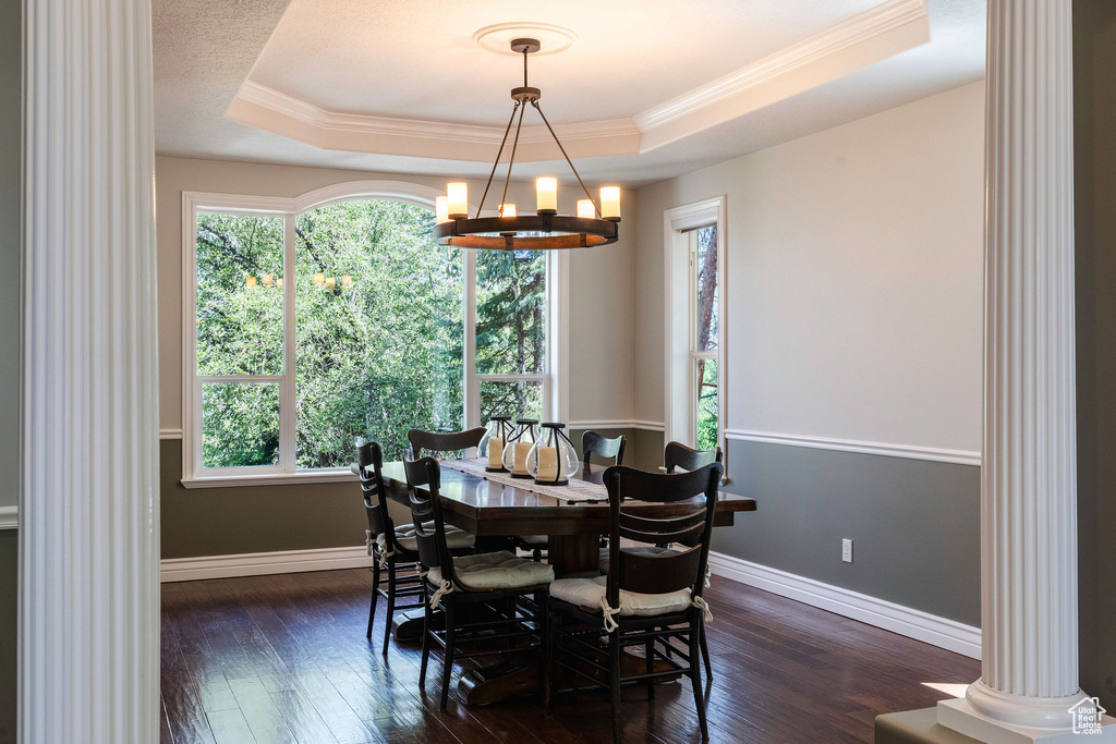 Dining space featuring a raised ceiling, decorative columns, a chandelier, ornamental molding, and dark wood-style flooring