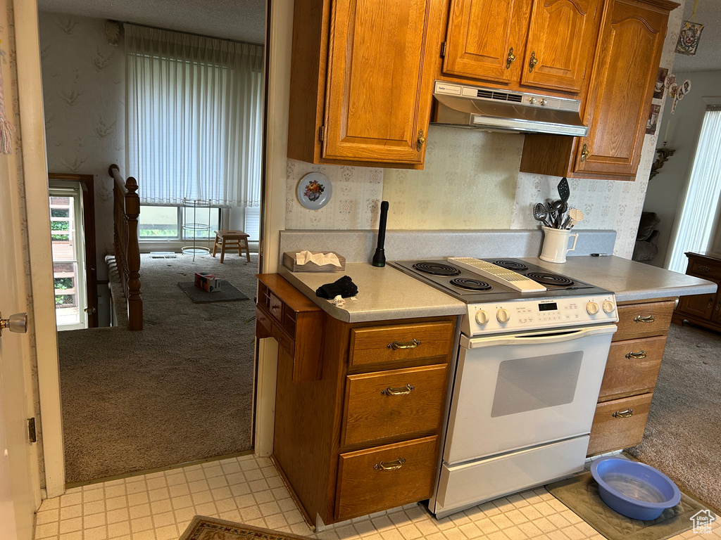 Kitchen with light colored carpet, wallpapered walls, brown cabinetry, under cabinet range hood, and white range with electric cooktop