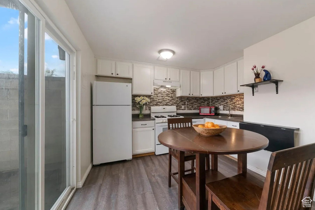 Kitchen with under cabinet range hood, dark countertops, white appliances, tasteful backsplash, and dark wood-style flooring