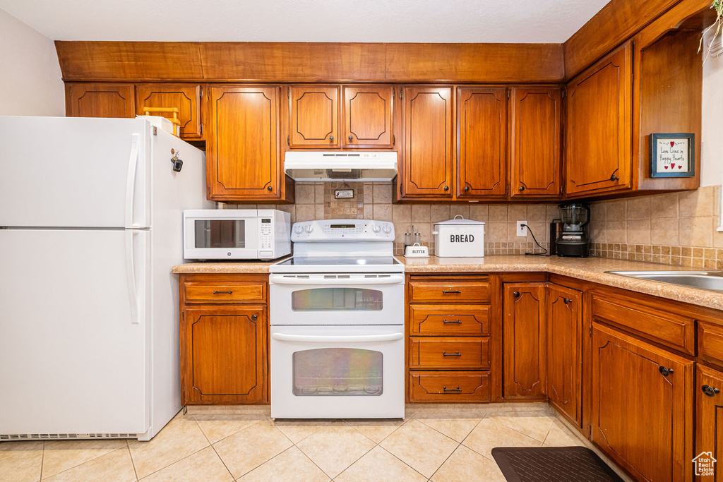 Kitchen with light tile patterned floors, brown cabinets, under cabinet range hood, and white appliances