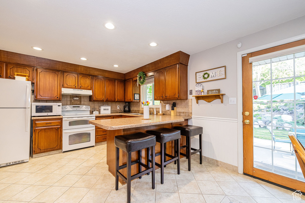 Kitchen featuring backsplash, light countertops, a peninsula, under cabinet range hood, and white appliances