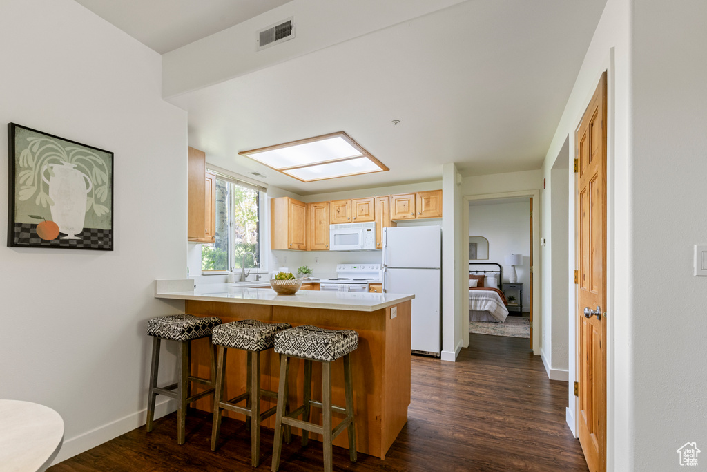 Kitchen with white appliances, a peninsula, dark wood finished floors, a kitchen bar, and light brown cabinets