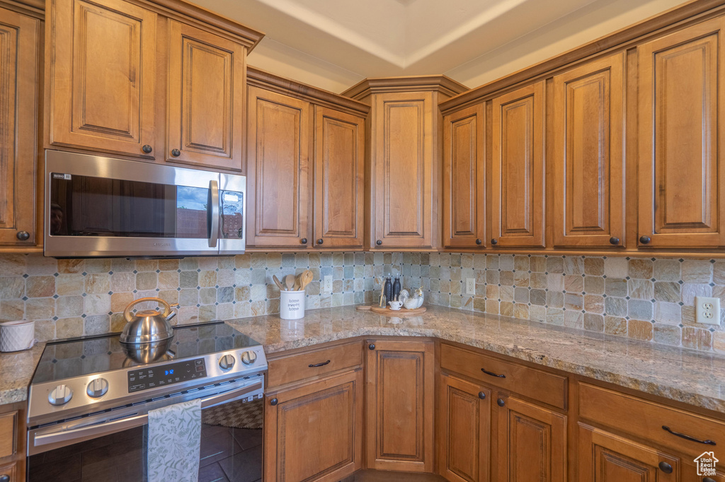 Kitchen featuring brown cabinetry, backsplash, appliances with stainless steel finishes, and light stone counters