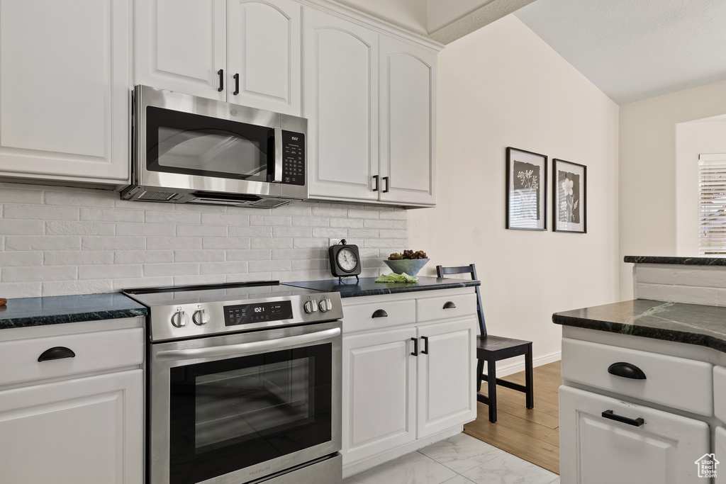 Kitchen featuring appliances with stainless steel finishes, backsplash, white cabinets, light marble finish floors, and dark stone counters