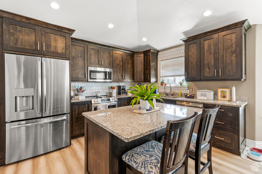 Kitchen featuring light wood finished floors, stainless steel appliances, a sink, dark brown cabinetry, and light stone counters