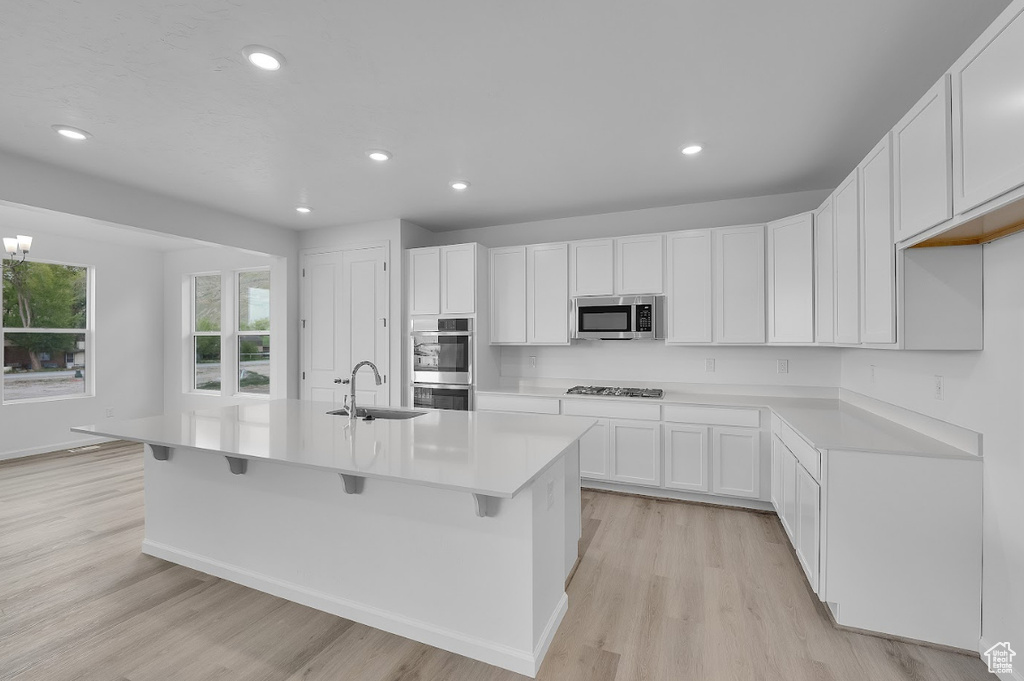 Kitchen with appliances with stainless steel finishes, a sink, light wood finished floors, an island with sink, and white cabinets