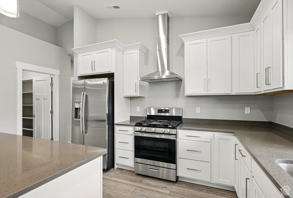 Kitchen with appliances with stainless steel finishes, wall chimney range hood, white cabinetry, vaulted ceiling, and light wood-type flooring
