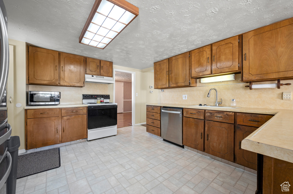 Kitchen with brown cabinetry, appliances with stainless steel finishes, a sink, light countertops, and under cabinet range hood