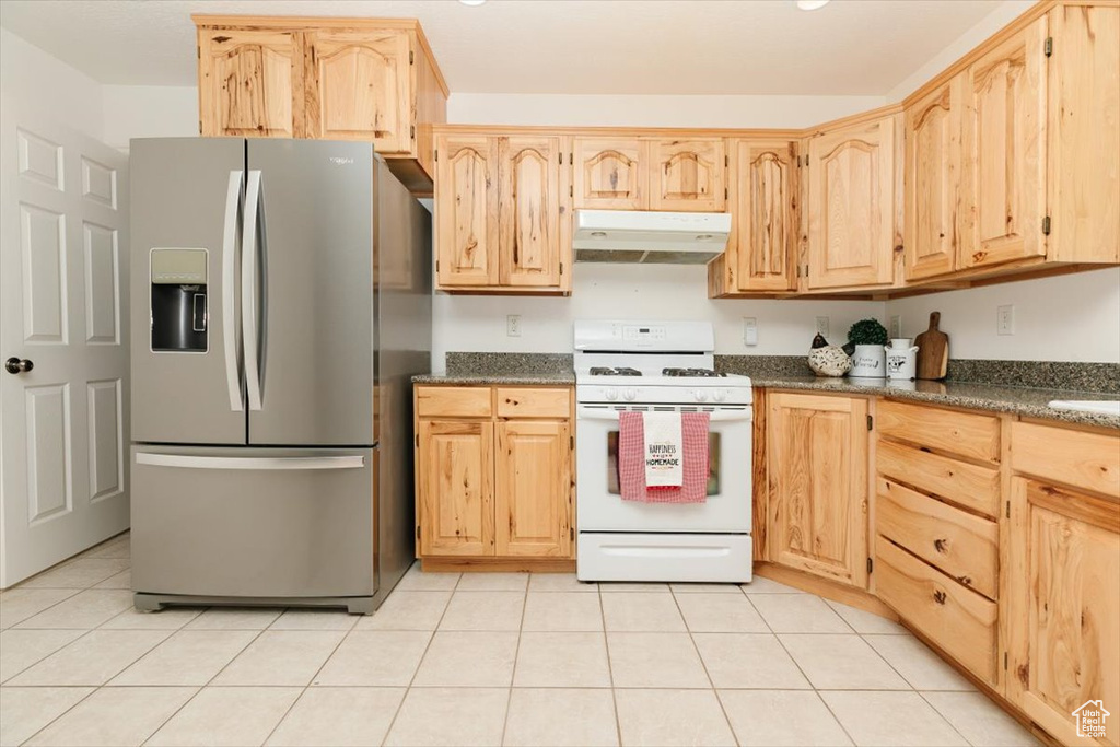 Kitchen with stainless steel fridge, white gas range, under cabinet range hood, light brown cabinets, and light tile patterned flooring