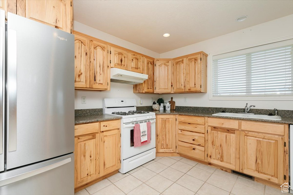 Kitchen with freestanding refrigerator, white gas stove, under cabinet range hood, light brown cabinets, and dark countertops