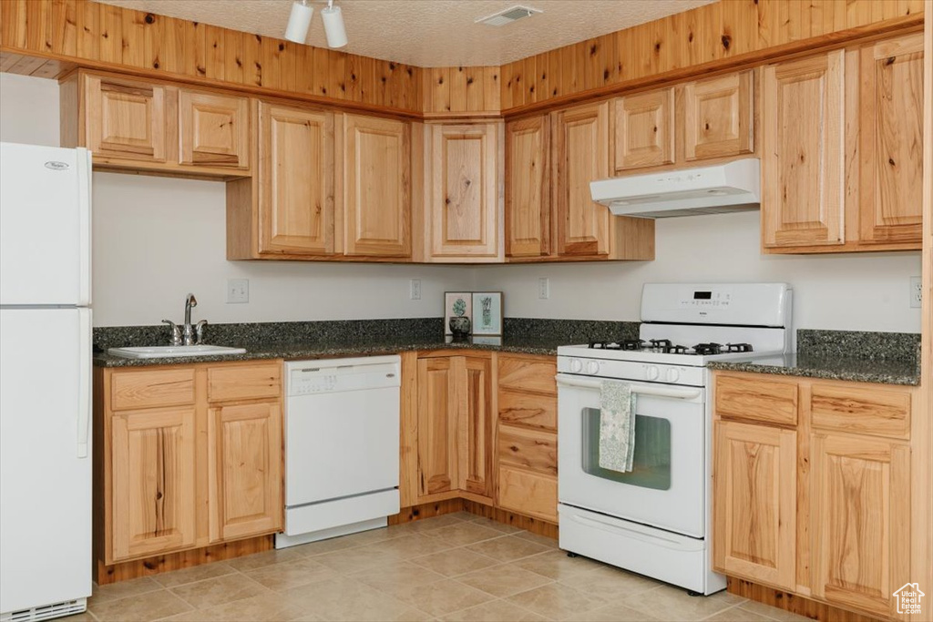Kitchen featuring white appliances, under cabinet range hood, dark stone counters, and light brown cabinets