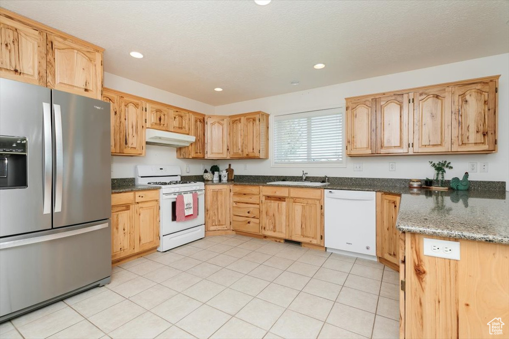 Kitchen with white appliances, under cabinet range hood, light brown cabinets, dark stone countertops, and recessed lighting