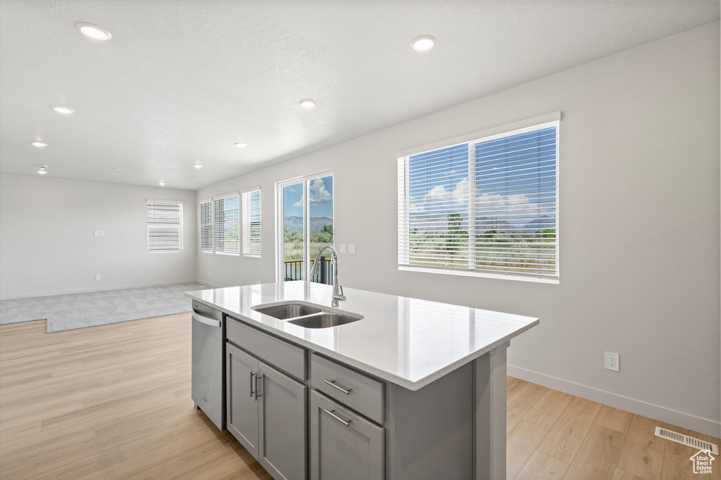 Kitchen featuring gray cabinets, stainless steel dishwasher, light wood-style flooring, light countertops, and a kitchen island with sink