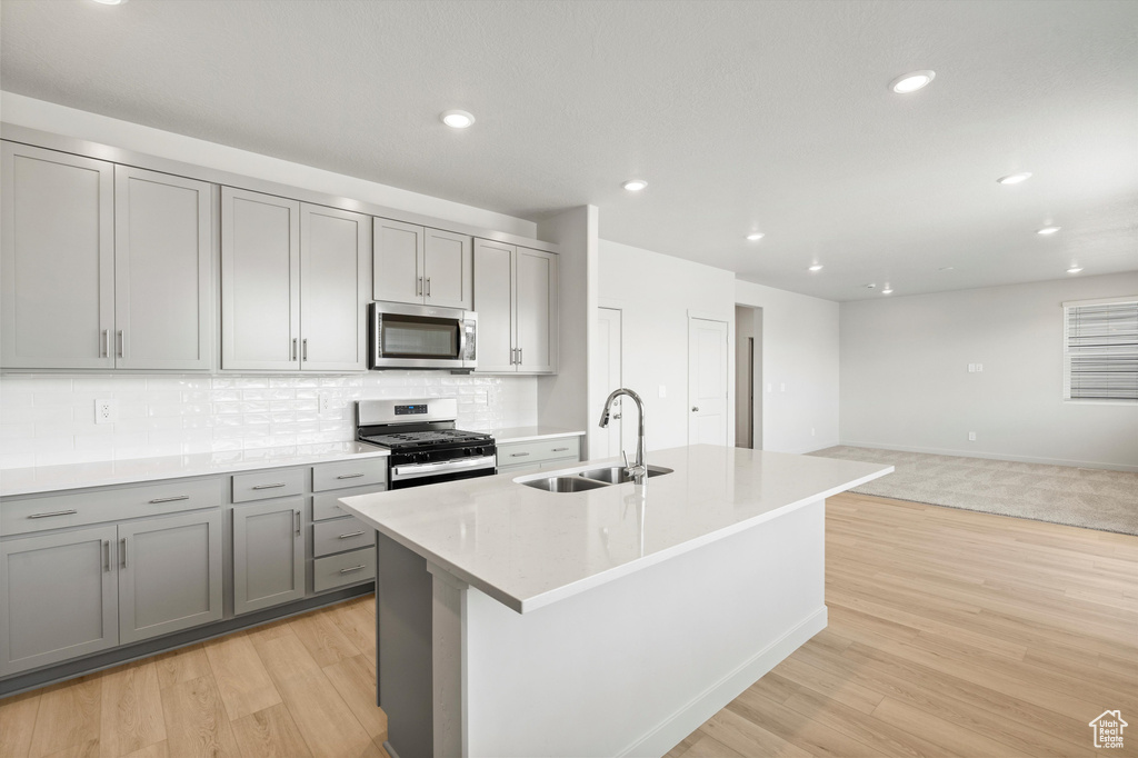 Kitchen featuring appliances with stainless steel finishes, gray cabinets, backsplash, light wood-style floors, and a center island with sink