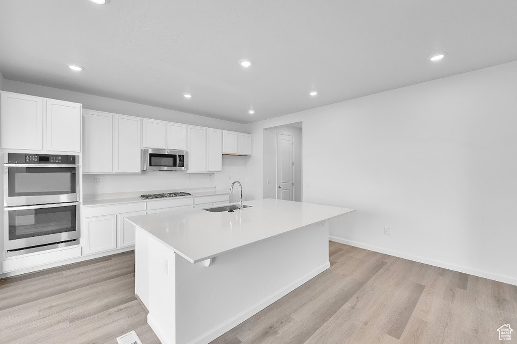 Kitchen featuring appliances with stainless steel finishes, a sink, white cabinetry, a center island with sink, and light wood-style floors