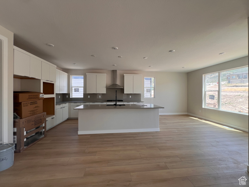 Kitchen featuring wall chimney exhaust hood, a kitchen island with sink, white cabinetry, light wood-type flooring, and recessed lighting