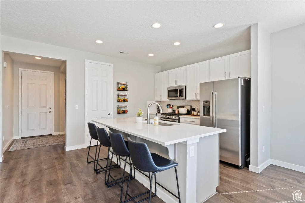 Kitchen with a sink, an island with sink, stainless steel appliances, white cabinets, and dark wood-style flooring