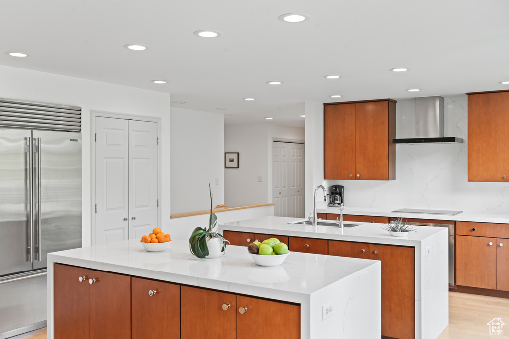 Kitchen featuring wall chimney range hood, built in fridge, a sink, recessed lighting, and an island with sink